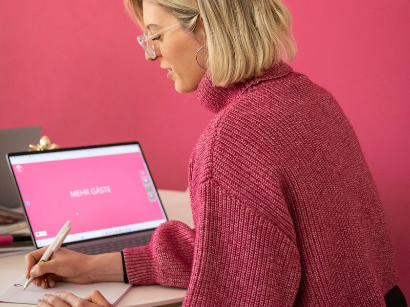 Frau mit blonden, kurzen Haaren, die in einem modernen Büro am Computer arbeitet und Notizen macht, vor einem rosa Hintergrund.