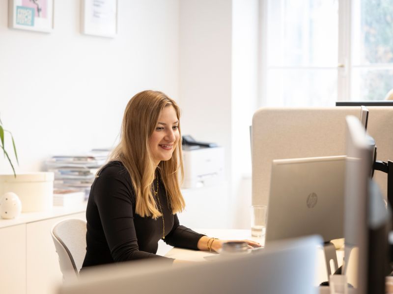 Eine junge Frau arbeitet lächelnd an einem Computer in einem modernen Büro, ideal für kreative Arbeitsumgebungen.