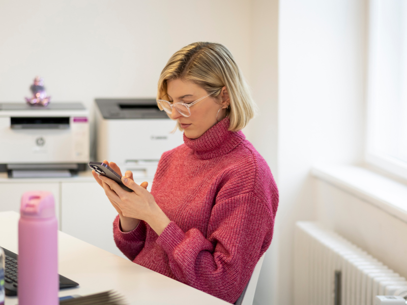 Eine Frau in einem modern gestalteten Büro sitzt an einem Tisch und nutzt ihr Smartphone, um Informationen zu überprüfen.