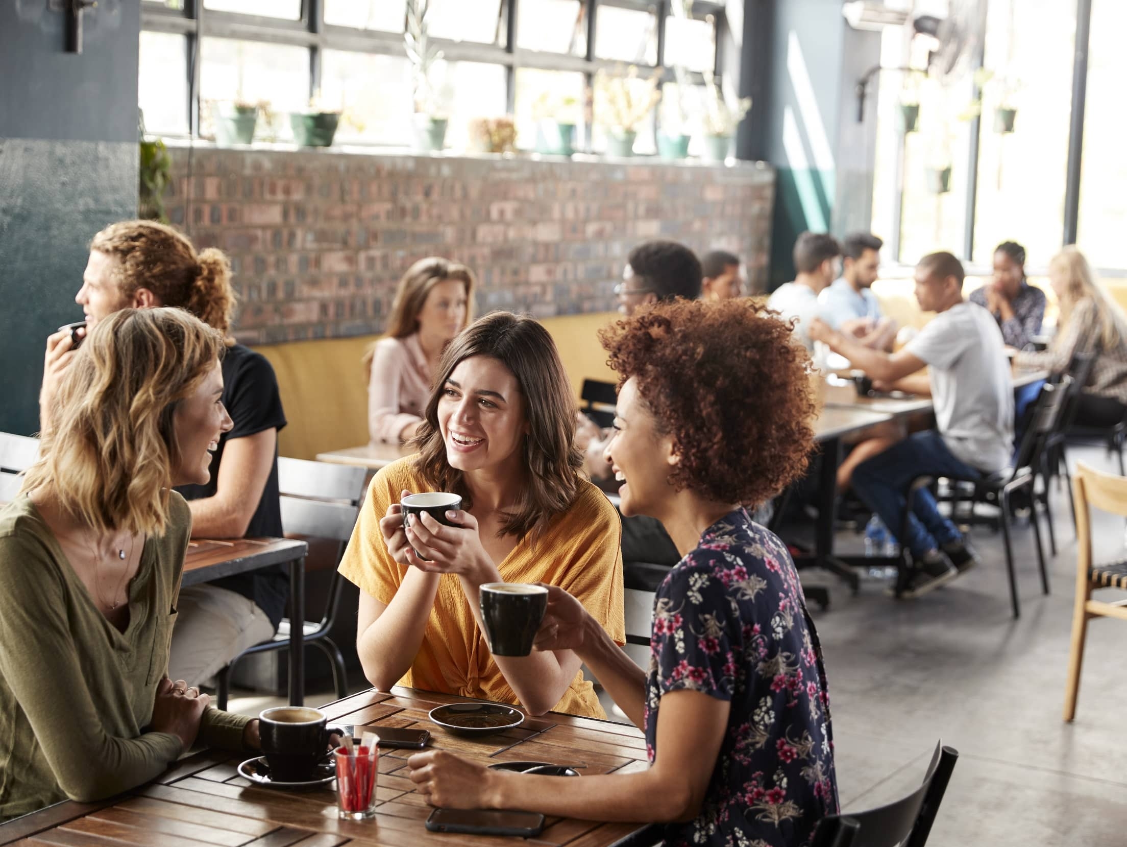 Freundinnen genießen Gespräche bei Kaffee in einem modernen Café, umgeben von anderen Gästen in entspannter Atmosphäre.