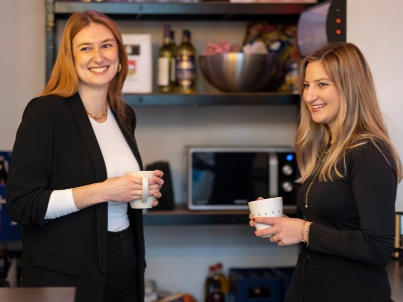 Zwei lächelnde Frauen stehen im Büro und unterhalten sich bei Kaffee, umgeben von modernen Büro-Utensilien und Geräten.