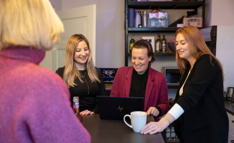 Drei Frauen arbeiten in einem modernen Büro zusammen an einem Laptop, während eine weitere Person im Hintergrund zuhört.