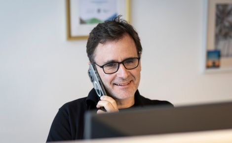 Ein Mann mit Brille spricht lächelnd am Telefon in einem modernen Büro, während er an einem Computer arbeitet.