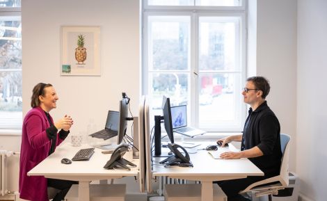 Zwei Kollegen im modernen Büro führen ein Gespräch, umgeben von Computern und Büromaterialien, ideal für den Arbeitsalltag.