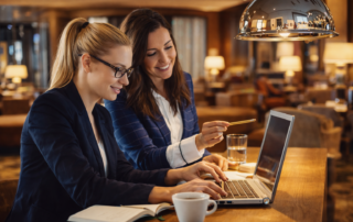 Zwei Frauen arbeiten gemeinsam an einem Laptop in einem stilvollen Café, umgeben von gemütlicher Atmosphäre und Getränken.