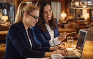 Zwei Frauen arbeiten gemeinsam an einem Laptop in einem stilvollen Café, umgeben von gemütlicher Atmosphäre und Getränken.