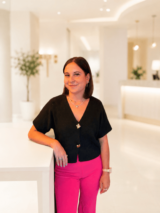 Freundliche Mitarbeiterin im modernen Hotel mit stilvollem Empfangsbereich Eine Mitarbeiterin steht im Empfangsbereich eines modernen Hotels, umgeben von eleganten Möbeln und Pflanzen, ideal für einen entspannten Aufenthalt.