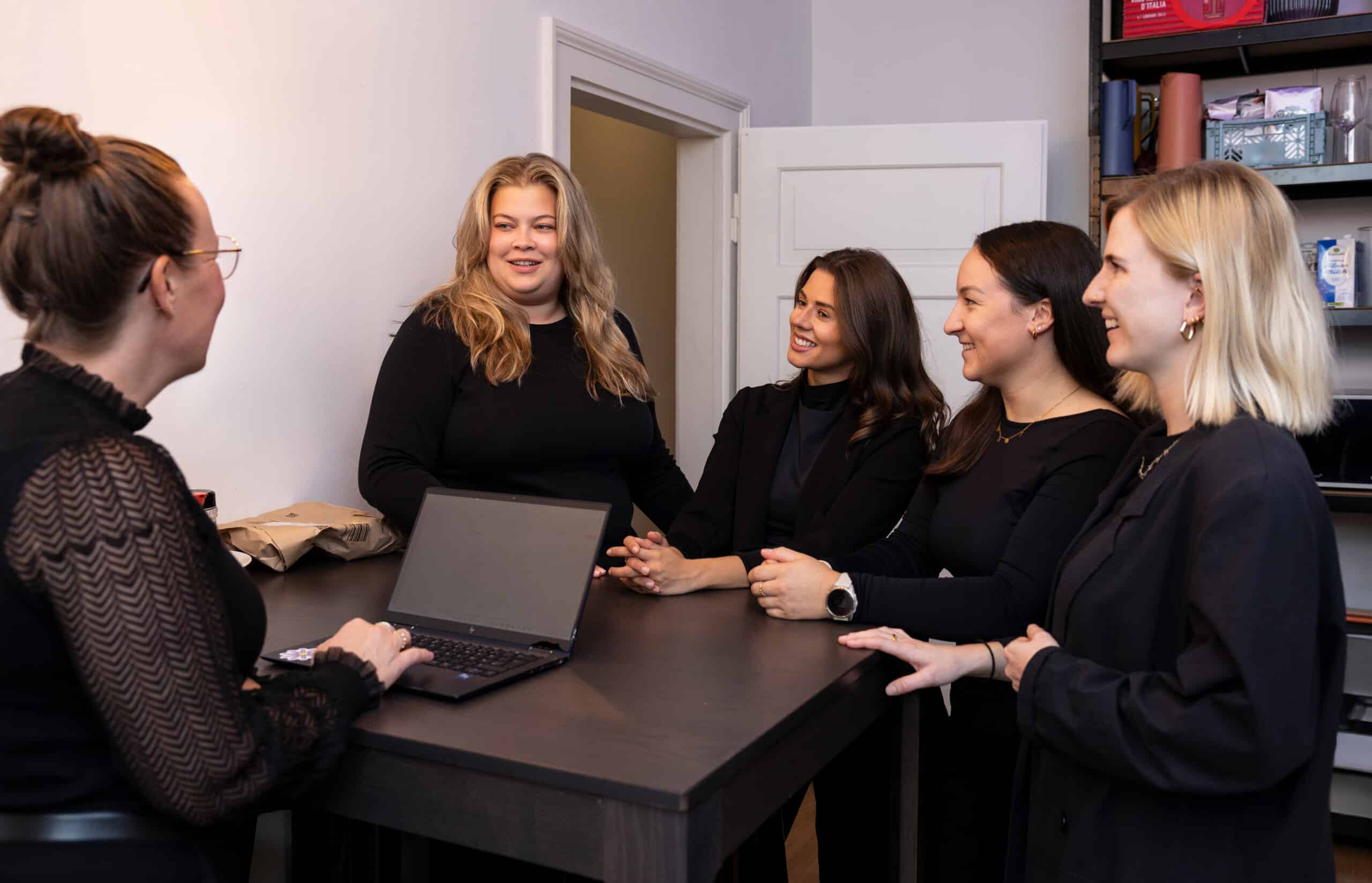 Vier Frauen in einem Büro während einer Teambesprechung, umgeben von einem modernen Arbeitsumfeld mit Laptop und Notizen.